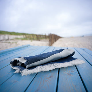 Blau-weißes Strandtuch auf blauem Holztisch, Dünen im Hintergrund – Teremana Merch